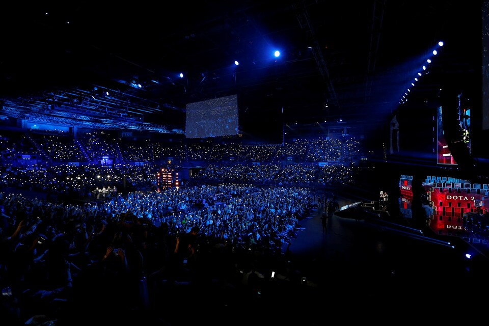 Une salle de spectacles est remplie de personnes. Les lumières sont tamisées bleues et tous les membres de la foule tiennent une petite lumière dans leur main.