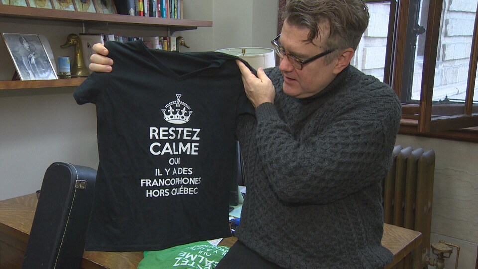 Dougald Lamont, assis sur le coin de son bureau, montre un T-shirt noir avec l'inscription « Restez calmes, oui il y a des francophones hors Québec ».