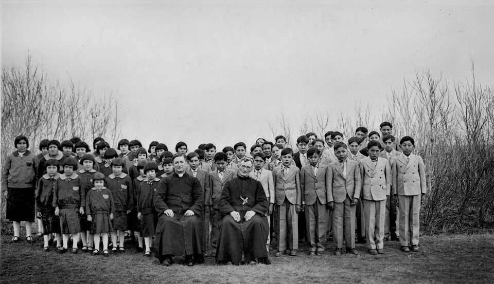 Des élèves du pensionnat autochtone Brocket, en Alberta, en 1930, sont debout en uniformes d'élèves derrière deux religieux assis.