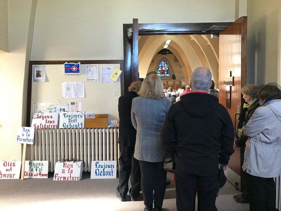 Quelques personnes observent la messe par les portes ouvertes, l'église étant déjà pleine. À leurs côtés, des affiches sont posées sur le sol, sur lesquelles on peut lire : Non à la fermeture.
