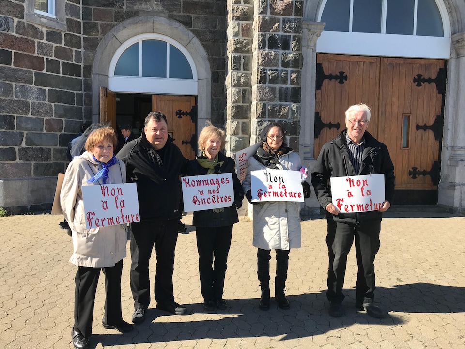 Cinq personnes se tiennent devant l'église avec des pancartes sur lesquelles on peut lire : Non à la fermeture.