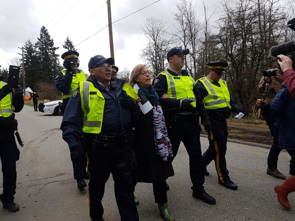 Une dame au bras de deux policiers et entourée de trois autres est escortée dans une rue, près d'une forêt, face aux caméras de télévision.