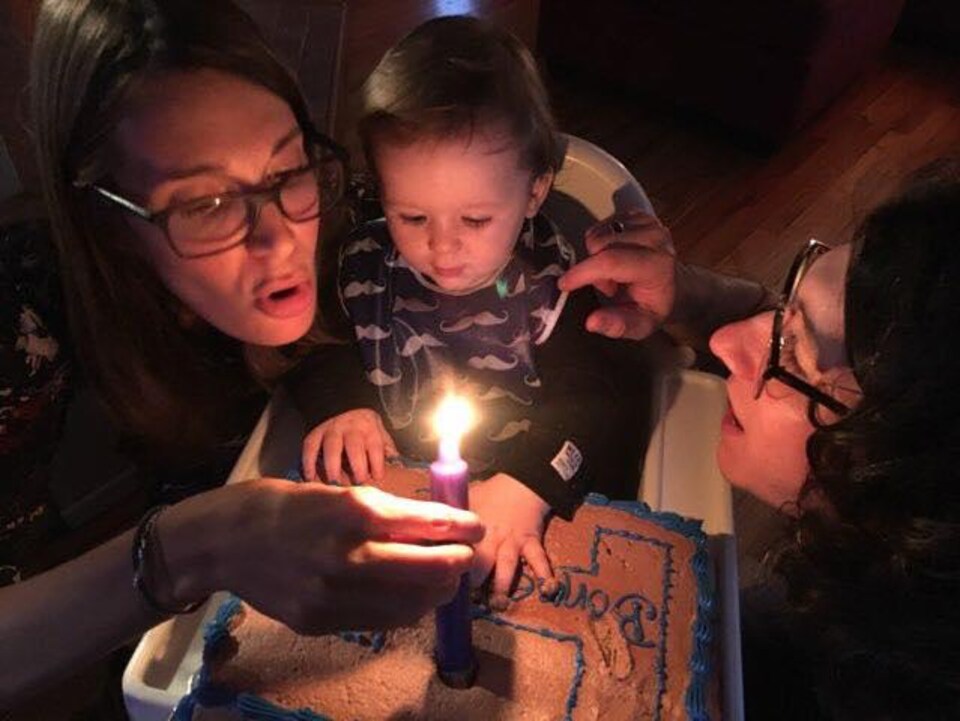 Un enfant sur une chaise devant un gâteau d'anniveraire et une bougie allumée avec ses parents de chaque côté.