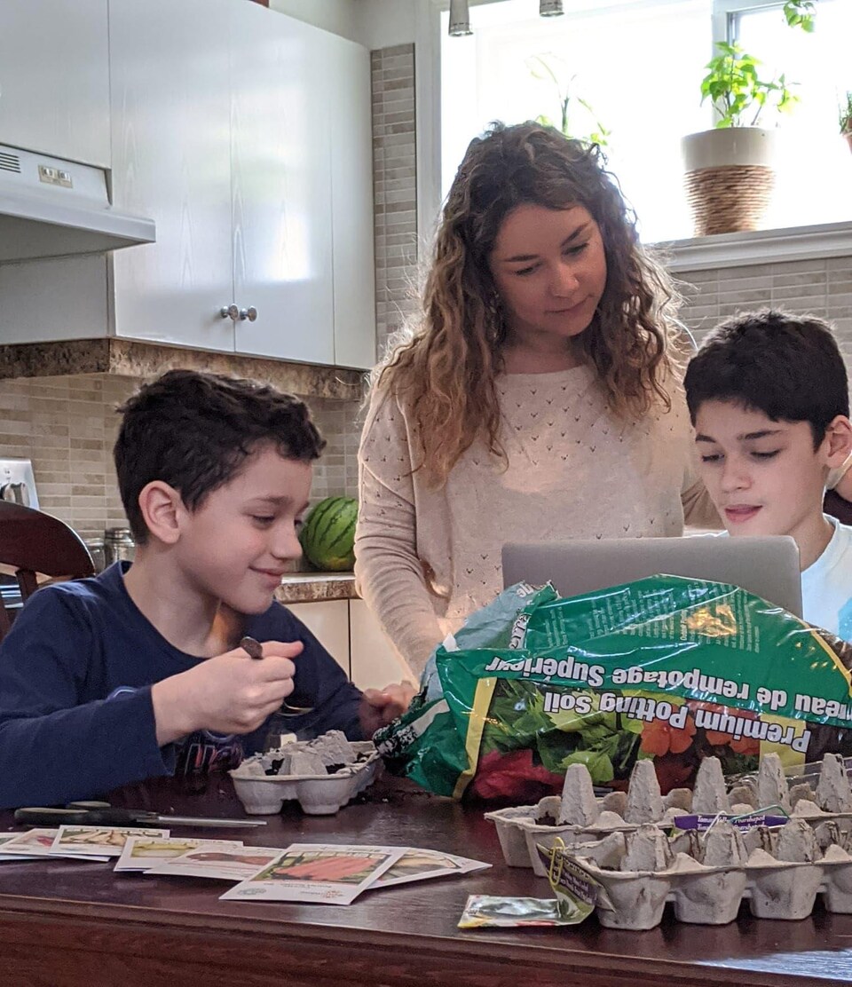 Caroline et ses enfants, devant un sac de terre, des boîtes à oeufs et des sachets de graines de légumes.