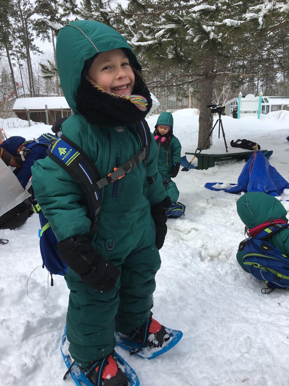 Un enfant de quatre ans en habit de neige émeraude et en raquettes avec un grand sourire, entouré de ses camarades de classe, eux aussi en habit de neige vert émeraude.