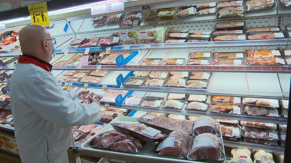 Un boucher, de dos, regarde le comptoir des viandes dans une épicerie.