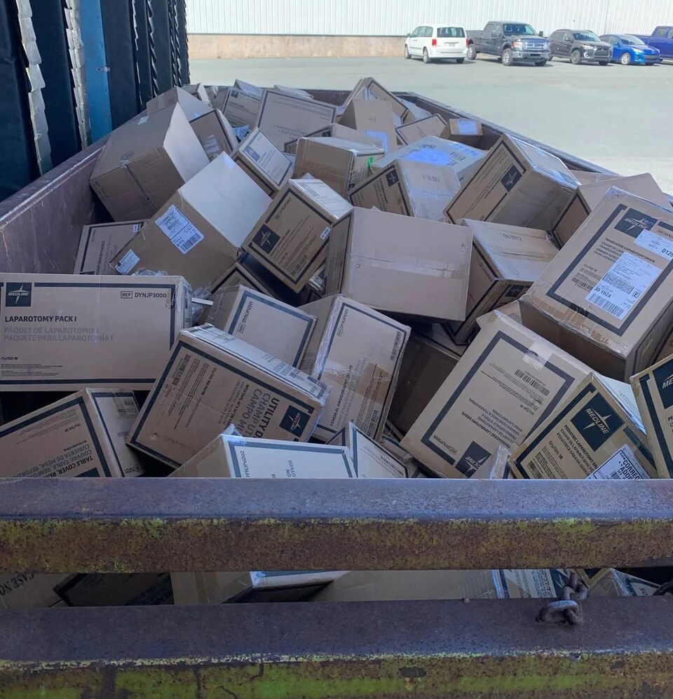 Des dizaines de boîtes jamais ouvertes et empilées dans un conteneur à déchets, dans un stationnement.