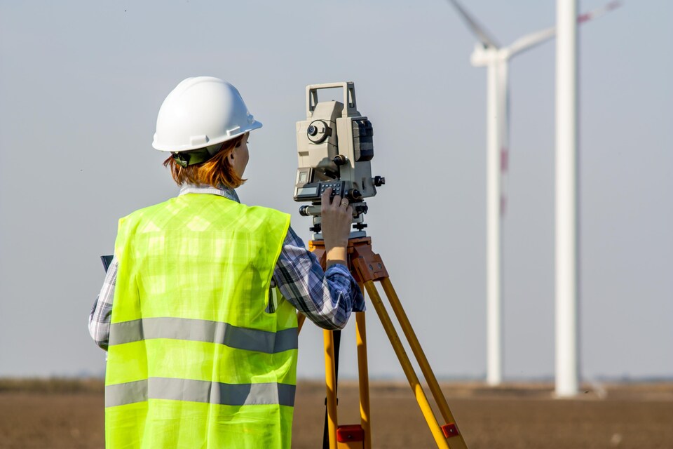 Une arpenteuse-géomètre dans un champ fait des mesures devant une éolienne.