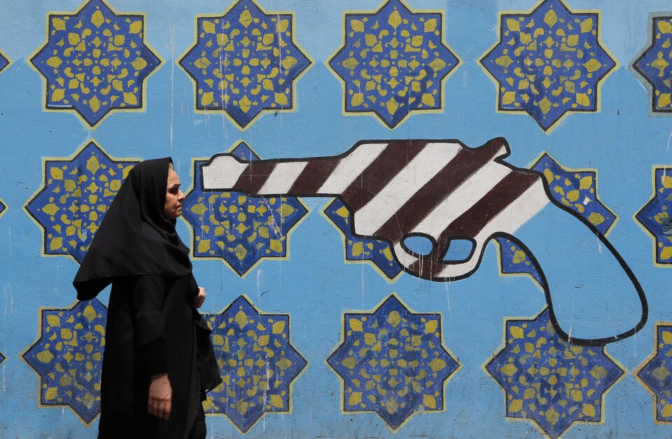 Une Iranienne marche devant une murale où l'on voit un pistolet peint aux couleurs du drapeau américain. 