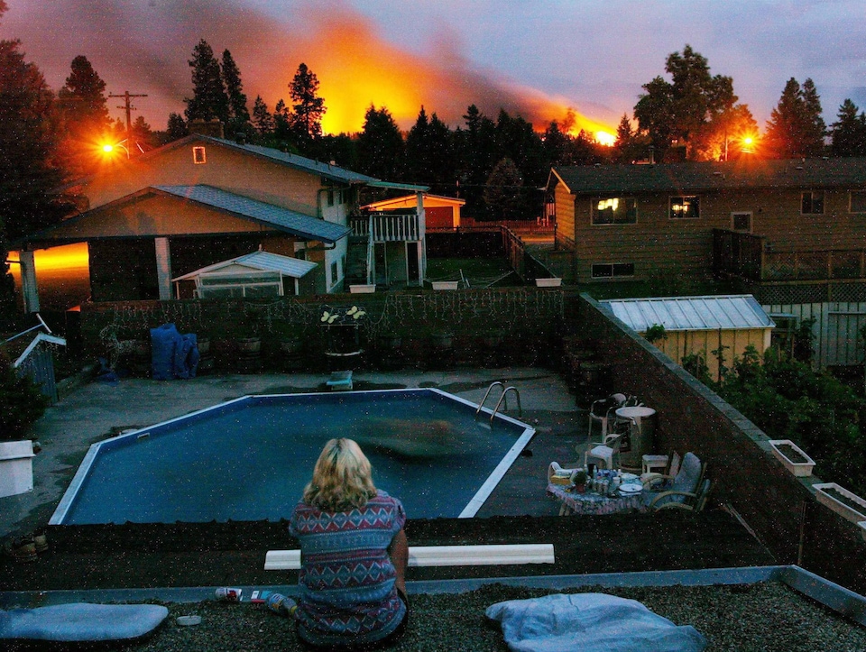 Une dame est assise face à une cour et des maisons avec, en arrière-plan, les lueurs d'un incendie.