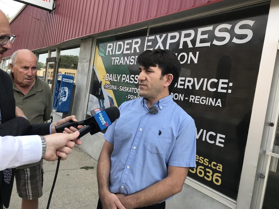 Le président de Rider Express Transportation, Firat Uray, se trouve devant la vitrine de son entreprise, à Regina. Il a les cheveux noirs et porte une chemise bleue. Deux journalistes sont devant lui et tendent leur micro.