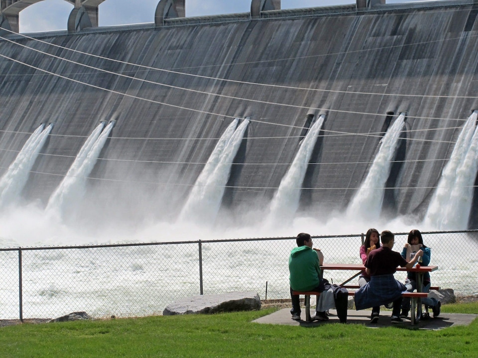 Des étudiants mangent à une table, devant l'imposant barrage Grand Coulee, sur le fleuve Columbia. Les vannes laissent échapper une grande quantité d'eau.