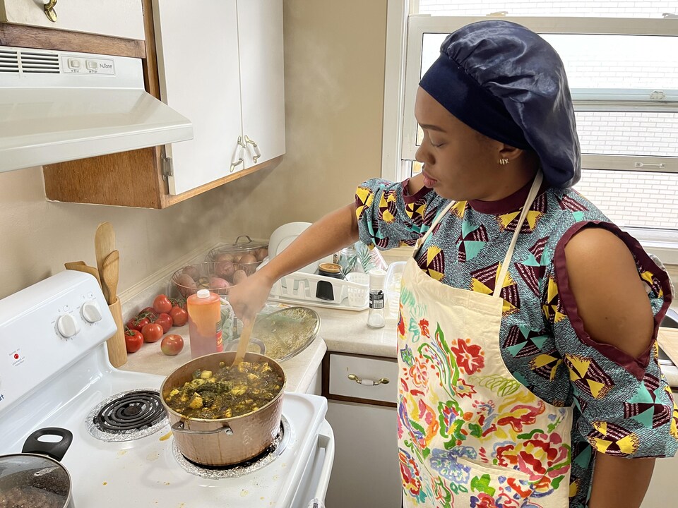 Une femme devant une cuisinière brasse quelque chose qui cuit dans un chaudron.