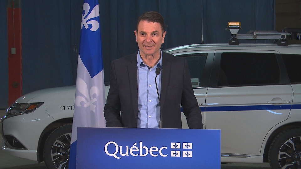 François Bonnardel au podium devant une voiture du ministère des Transports et un drapeau du Québec