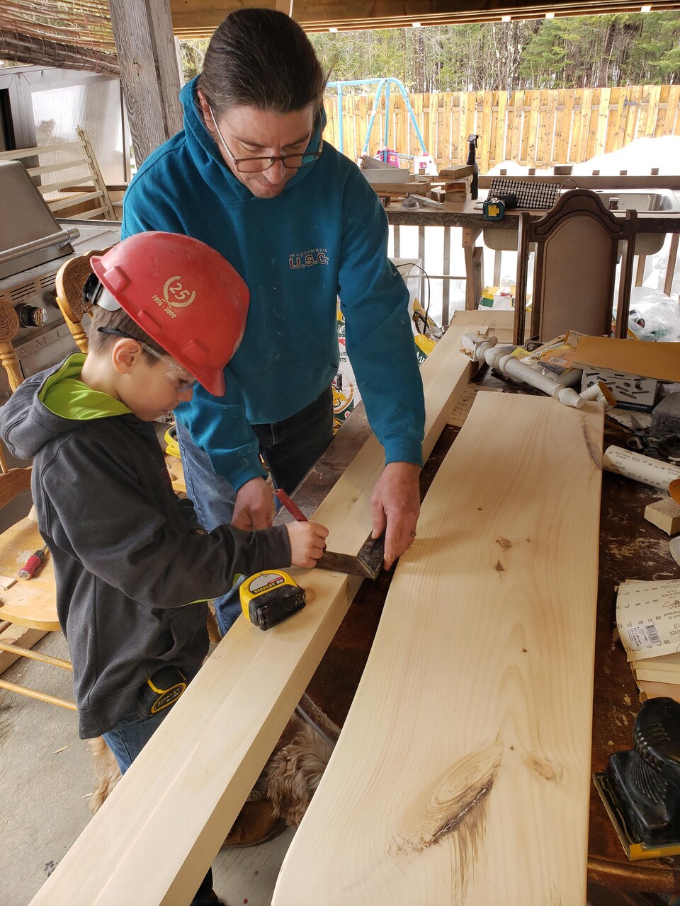 Le père et son enfant devant des planches de bois. Le petit mesure une planche.