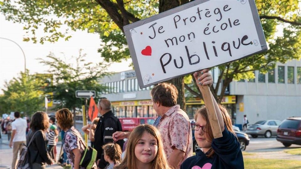 Une jeune fille tient une pancarte sur laquelle est écrit : « Je protège mon école publique ». On peut voir de nombreuses personnes former une chaîne humaine en arrière-plan.
