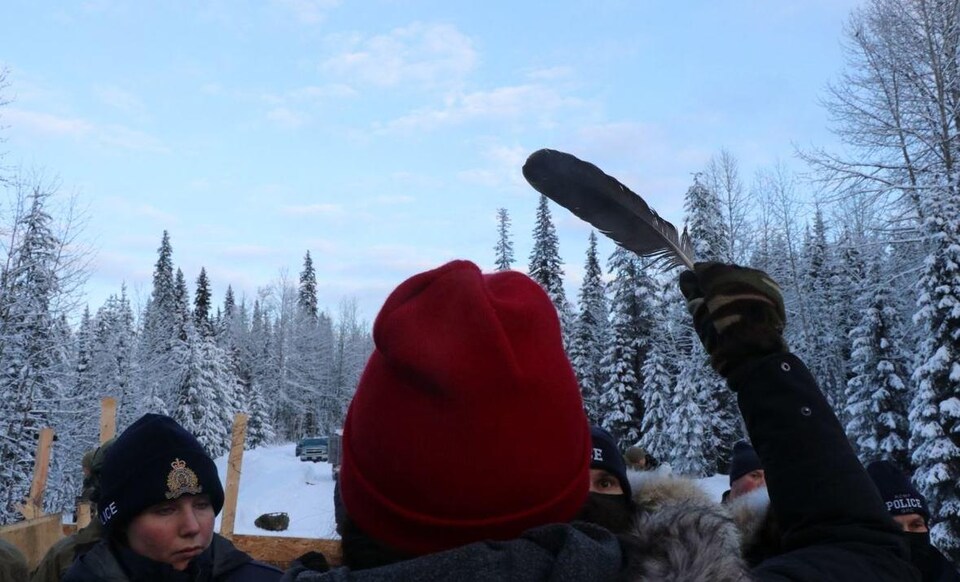 Un manifestant tient une plume dans la main droite devant des policiers dans une forêt recouverte de neige. 