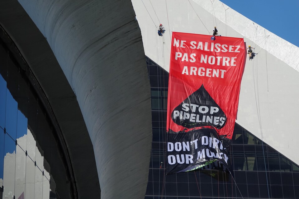 Des personnes, munies de câbles et de harnais, ont déployé une longue banderole sur laquelle ont peut lire « Ne salissez pas notre argent : stop pipelines ». Ils suspendus le long d'une façade.