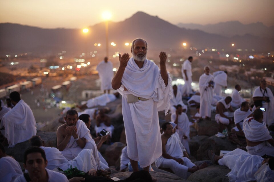 Vêtu d'un toge blanche, un pèlerin musulman prie au mont Arafat en levant les mains. Il est entouré de dizaine d'autres croyants.