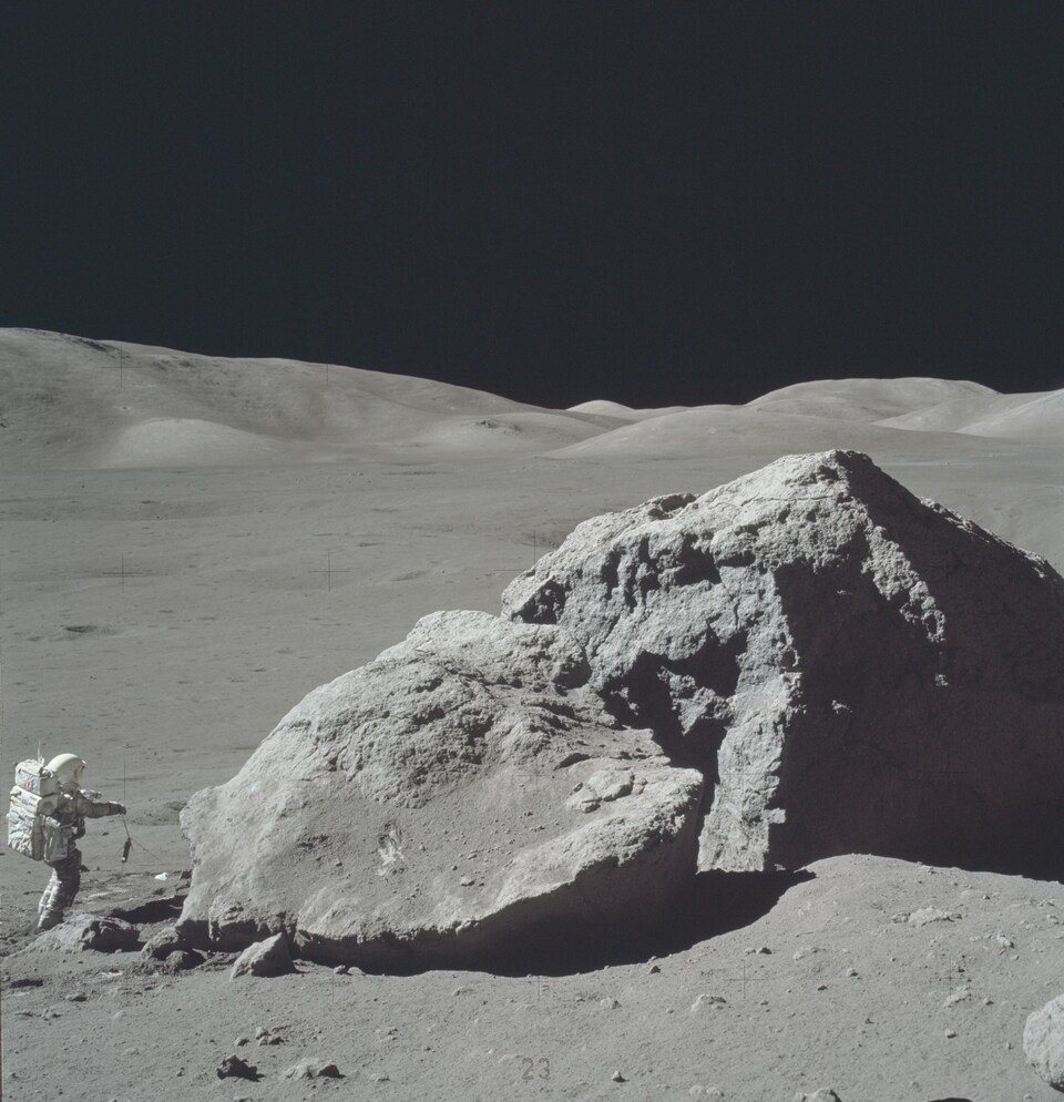 Une photo en noir et blanc d'un astronaute sur la Lune en 1972. Il semble tout petit et est debout face à un très gros rocher.