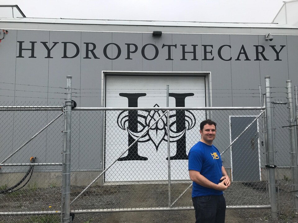 Un homme portant un t-shirt et des jeans sourit en regardant en notre direction. Derrière lui se trouve un logo, un « H », et un mot, soit « Hydrophothecary ».