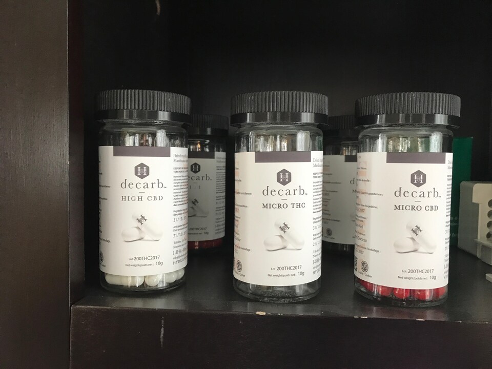 De petits pots contenant des pilules sont posés sur une tablette. Sur leur étiquette, on peut notamment lire « decarb » et voir le logo de la compagnie Hydropothicaire qui ressemble à un « H ».