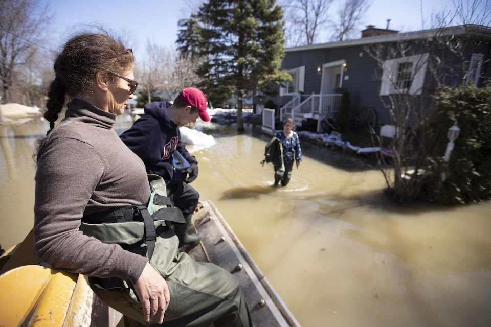 Une femme et un homme sont transportés dans une pelle de tracteur. Une femme devant une maison située tout près a les pieds dans l'eau.