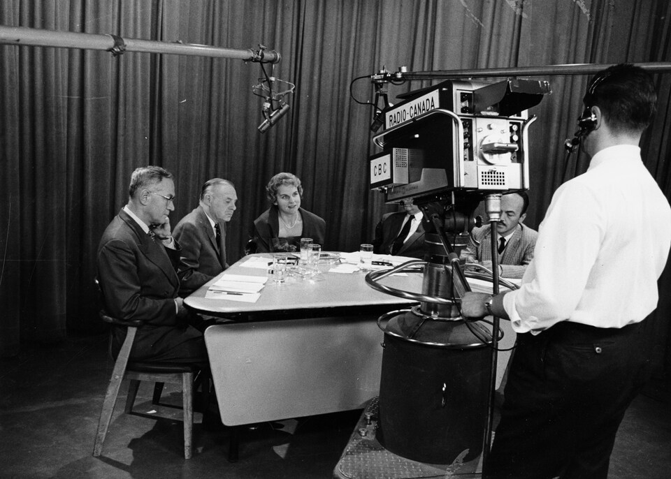 Dans un studio de télévision, réunis autour d'une table, Paul Sauriol, Roger Champoux, Jeanne Sauvé, Lucien Desbiens (caché derrière la caméra) et Réal Benoît. Dans le plan, un micro sur perche et un caméraman derrière sa caméra sur trépied.