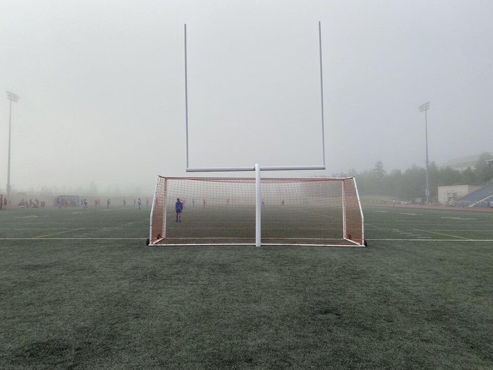 Un match de soccer qui se déroule dans le brouillard. La photo est prise derrière le but de soccer, au-dessus duquel se trouve le poteau des buts au football américain.