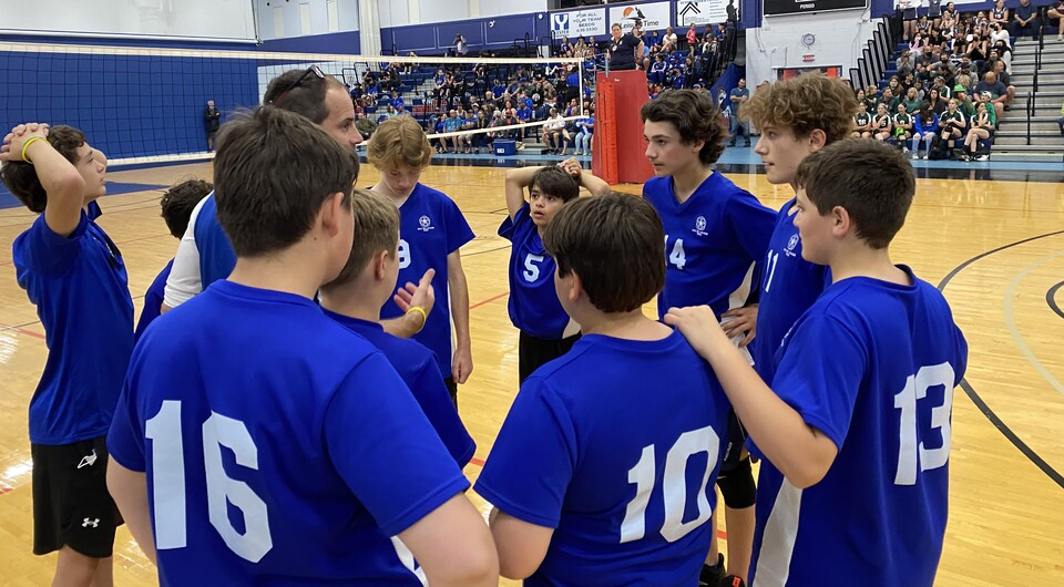 Des garçons qui jouent au volleyball font un caucus avec leur entraîneur sur les lignes de côté, dans un gymnase.