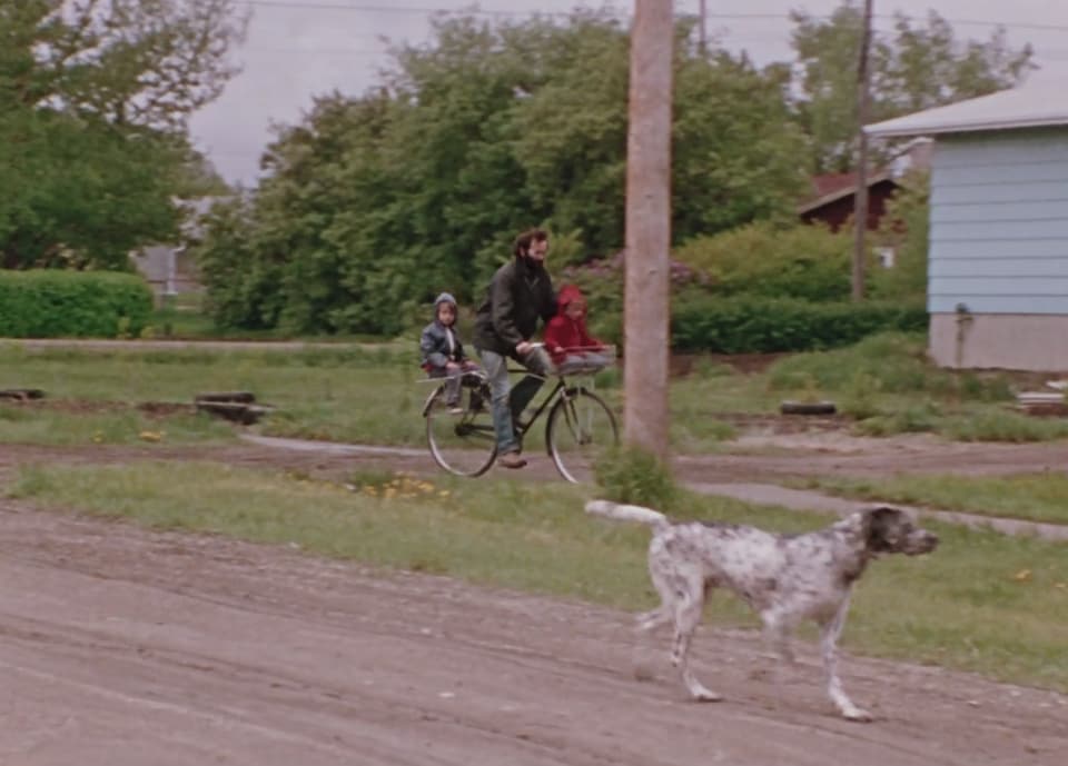 Joe Fafard sur son vélo avec deux enfants ainsi qu'un chien qui les suit. 