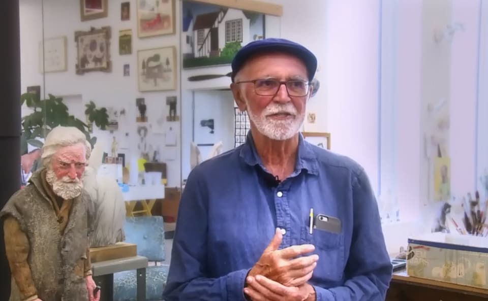 Joe Fafard se tient devant la caméra et porte un chapeau avec une chemise bleue. On voit derrière lui de nombreuses oeuvres d'art.