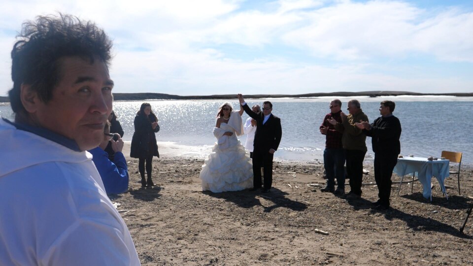 Un homme regarde la caméra et sourit, alors que, derrière lui, un mariage se tient devant l’océan Arctique.