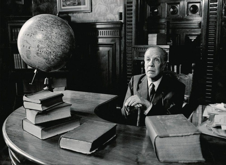 Portrait de Jorge Luis Borges assis sur un bureau en demi-cercle, dans une bibliothèque. 5 gros livres se trouvent sur le bureau.