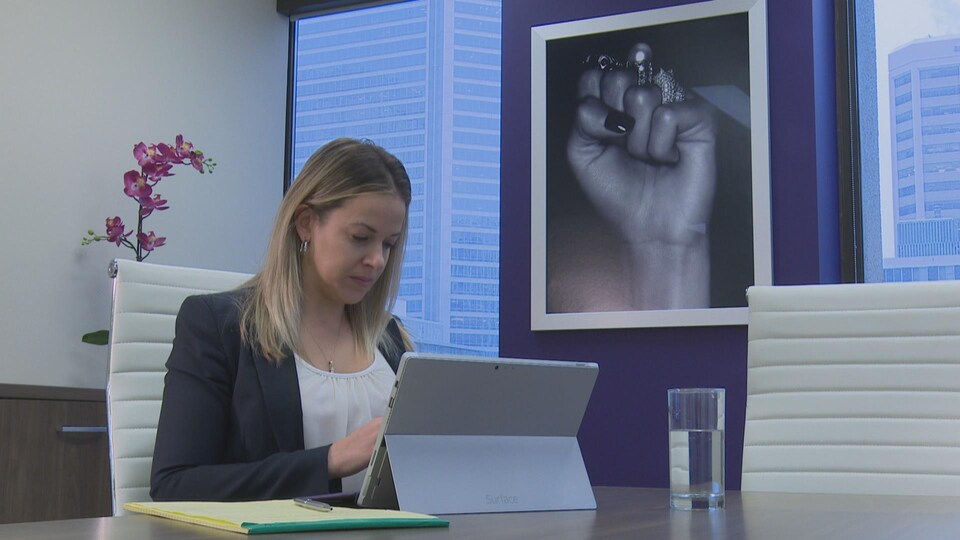 Une femme est assise à une table. Elle regarde son ordinateur. Un verre d'eau est posé sur la table. Derrière, on peut voir une plante ainsi qu'une photo représentant le poing fermé d'une femme.