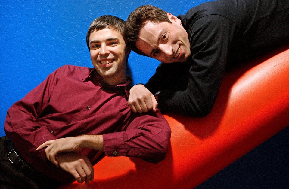 Une photo de Larry Page et de Sergey Brin, souriants, appuyés sur une sculpture tubulaire rouge, devant un fond bleu.