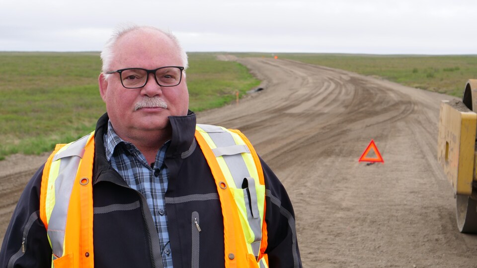 Un homme avec un dossard orange de construction se tient sur une route de gravier en construction.