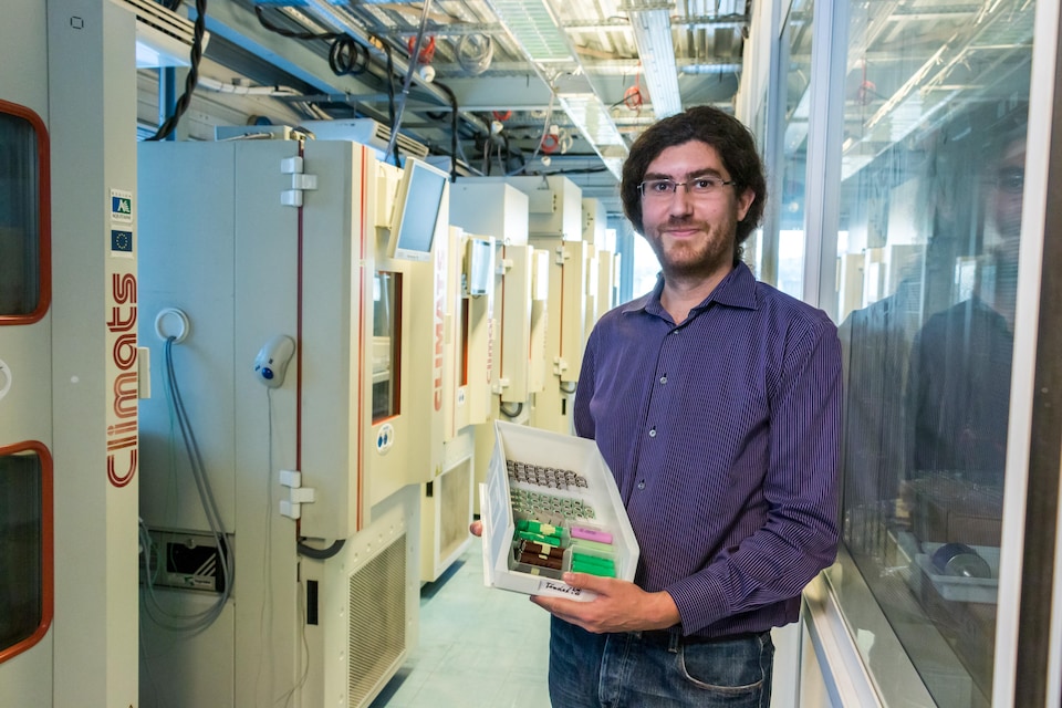 Loïc Boulon dans le laboratoire contenant des climatiseurs, tient une boîte de batteries dans les mains.