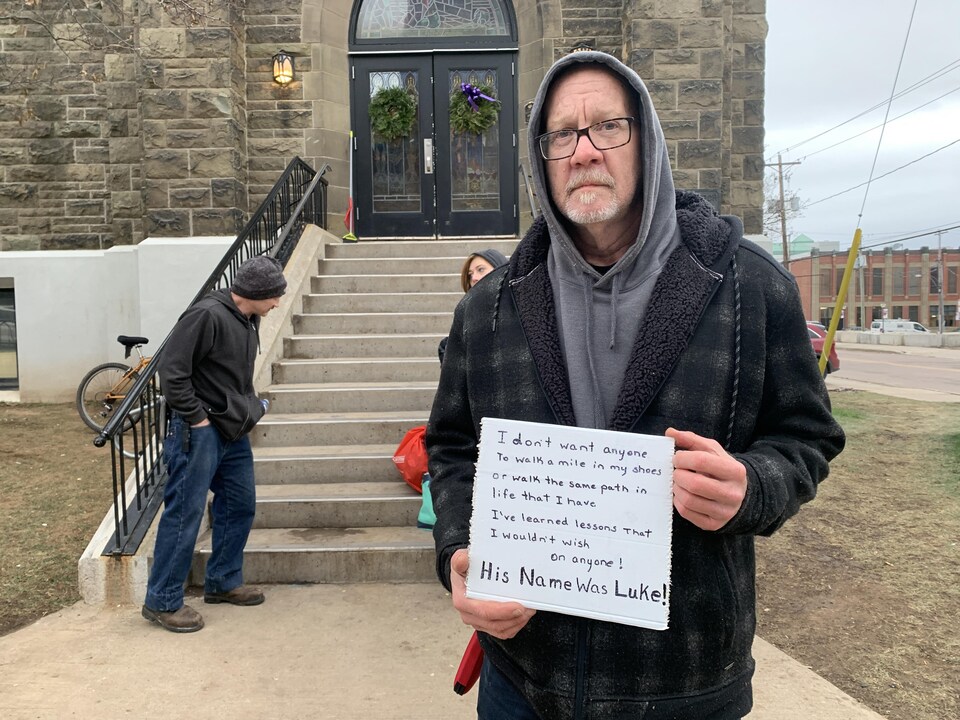 Un homme debout à l'extérieur d'une église tient un écriteau carré sur lequel est écrit un court poème, qui se conclut par la phrase « Son nom était Luke ».