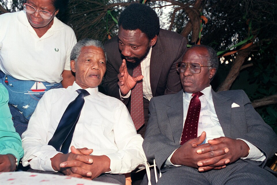 Cyril Ramaphosa debout, à l'arrière de Nelson Mandela et d'Andrew Mlangeni, qui sont assis, les mains croisées. 
