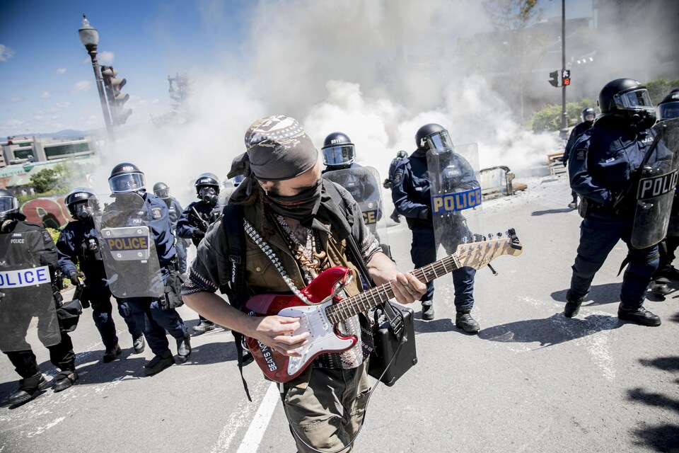 Un manifestant joue de la guitare électrique. Il transporte un amplificateur portatif en bandoulière. Son visage est en partie dissimulé par un foulard. En arrière de lui, des policiers antiémeutes équipés de boucliers et de matraques marchent en direction des manifestants. On aperçoit également un large panache de fumée.