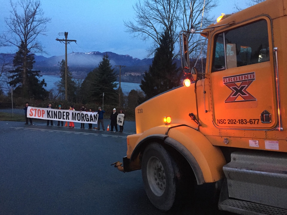 Des gens tiennent une banderole sur laquelle il est écrit : Stop Kinder Morgan en lettres majuscules. Ils se tiennent devant un immense camion.