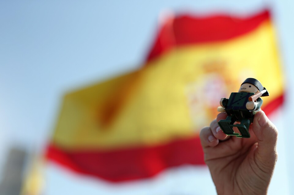 Une main tient une figurine d'un policier de la Garde civile espagnole pendant une manifestation. Hors-champ, on aperçoit un drapeau de l'Espagne.