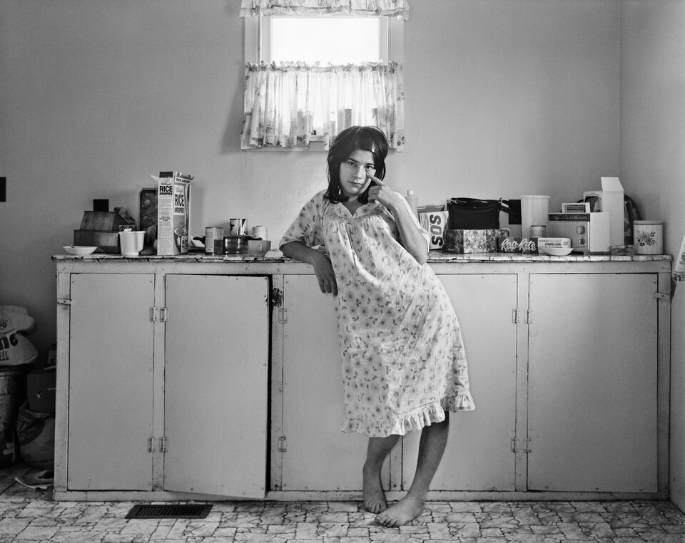 Photo en noir et blanc prise dans les années 1970 d'une jeune femme en jaquette, pieds nus, accoudée à un comptoir de cuisine couvert d'objets comme une boîte de céréales, une boîte de savon SOS et une radio. La femme étire la paupière inférieure de son œil gauche avec son index.