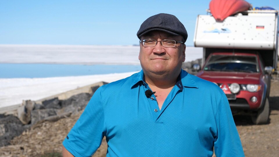 Un homme sourit pour la caméra, derrière lui, un véhicule récréatif et l’océan Arctique.