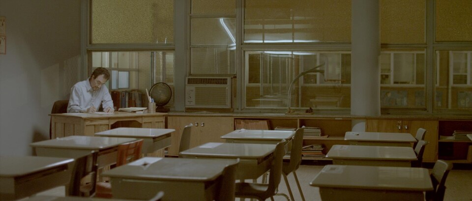 Une scène tirée du film « Monsieur Lazhar » où l'on voit l'acteur principal en train d'écrire seul, sur une table, assis dans une salle de classe