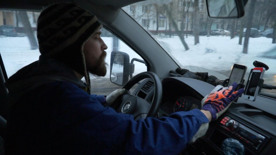 Un homme portant une tuque manipule son téléphone tout en conduisant une voiture dans une rue enneigée.