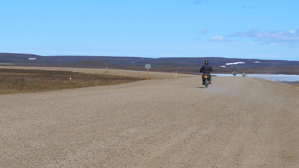 Quatre motocyclistes roulent au loin sur une route de gravier, avec la toundra et un lac encore gelé en arrière-plan.