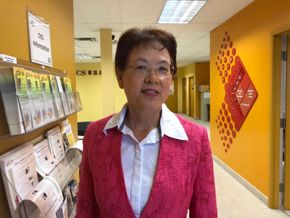 Une femme asiatique est debout dans un corridor à côté d'une étagère de dépliants. Elle porte des lunettes rondes, un cardigan et une chemise.