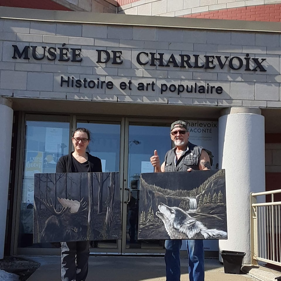 Daniel Bonneau et Nadyne Deschênes posent devant le Musée de Charlevoix portant chacun un tableau. 
